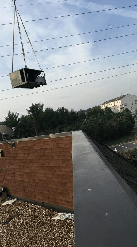 A crane suspends an HVAC unit in the air above a brick building roof on a sunny day.