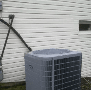 A grey HVAC condenser unit sits against a white-sided house wall with exposed black piping and a small window nearby.