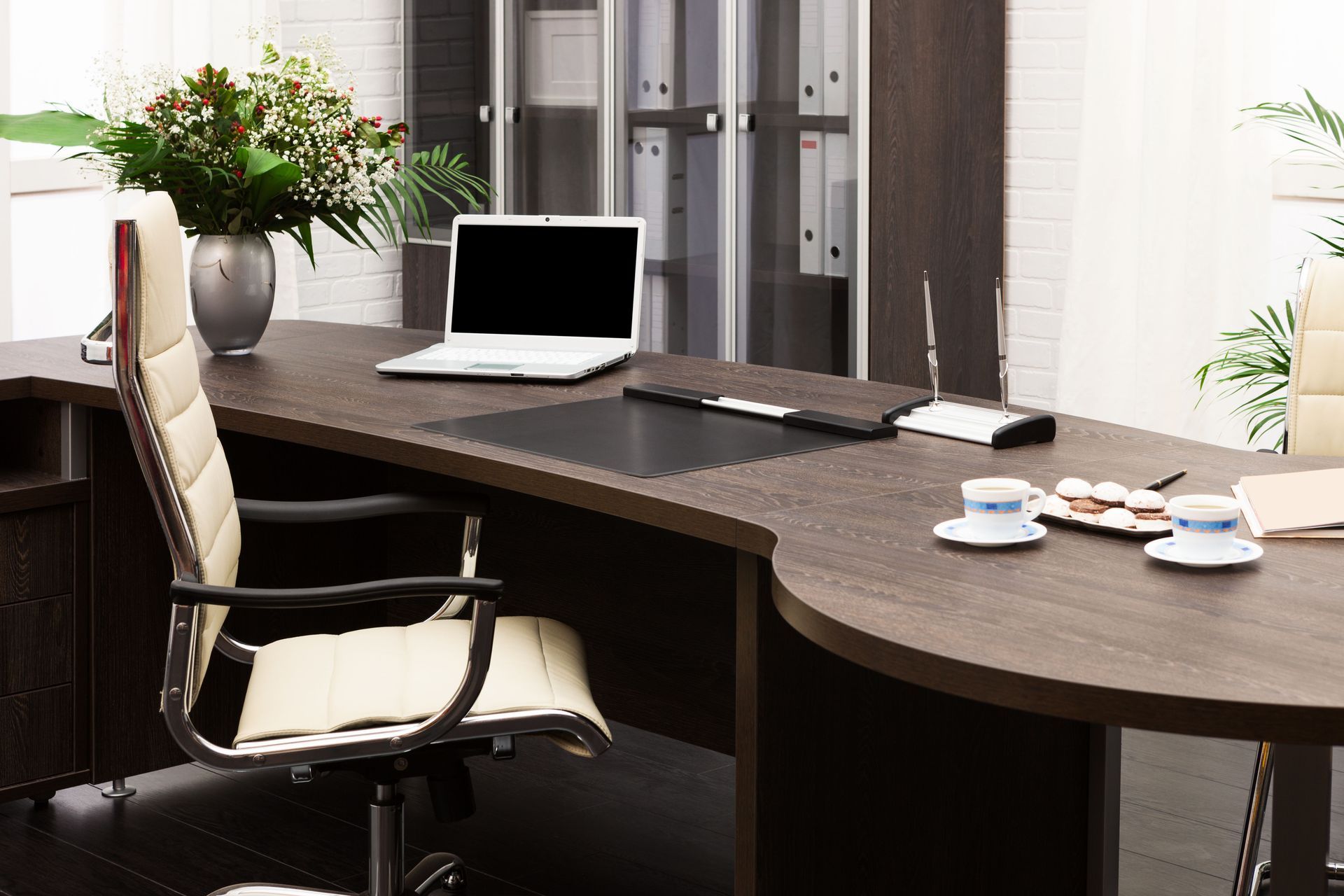Office desk with laptop, beige chair, flowers, and cups. Dark wood furniture in a bright room.