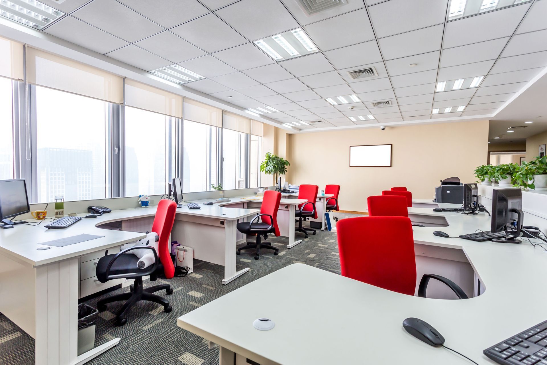 Modern office workspace with desks, chairs, computers, and windows. Red and black chairs, bright overhead lighting.