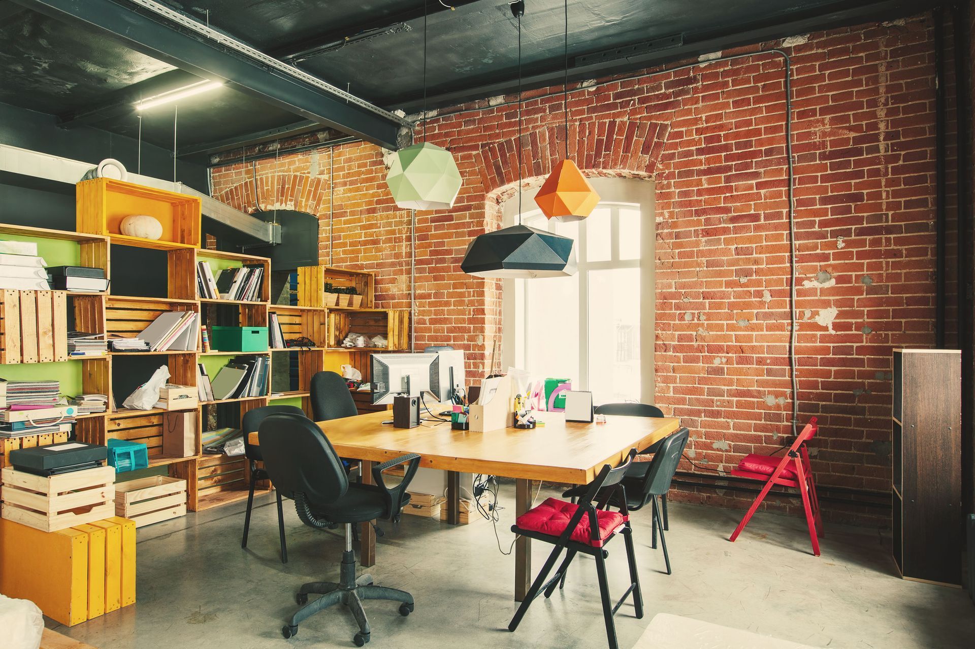 Office with exposed brick wall, wooden shelves, a table, and geometric pendant lights.