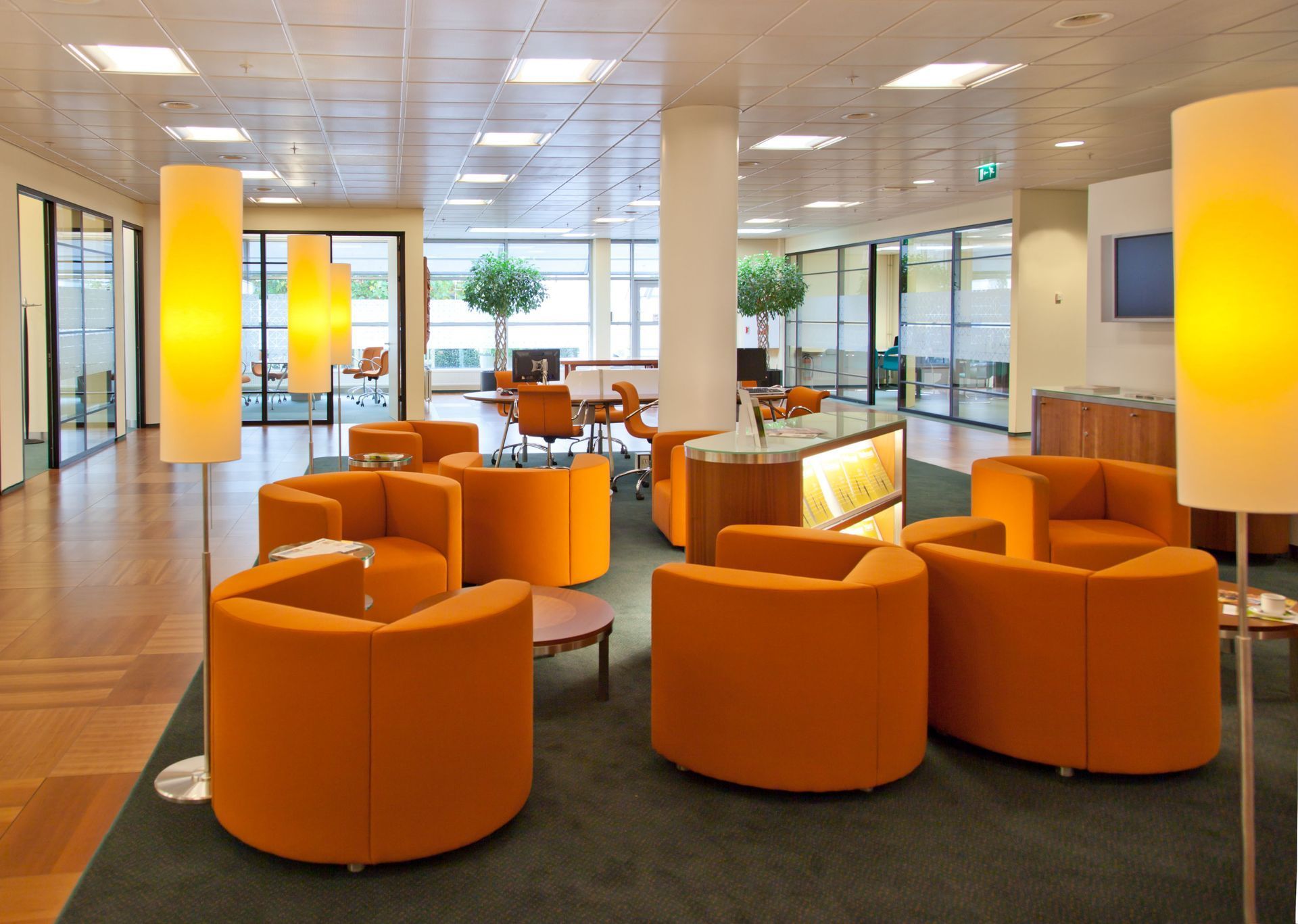 Lounge area with orange circular chairs, floor lamps, and a view of a light-filled office space.