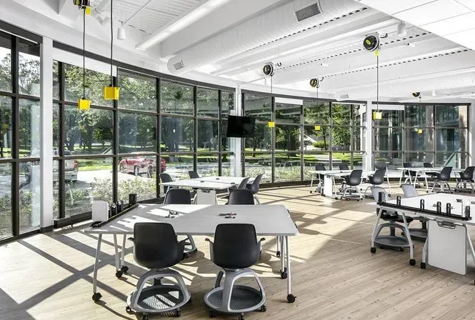 Classroom with large windows, tables, and swivel chairs. Natural light illuminates the room.
