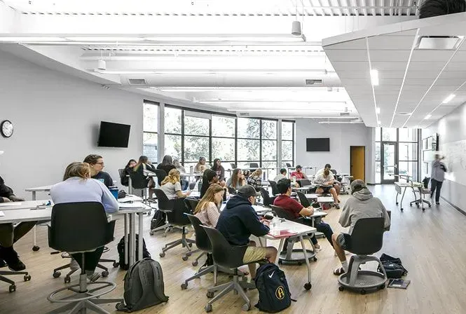 Classroom with students seated at tables, some facing a whiteboard where a person is writing.