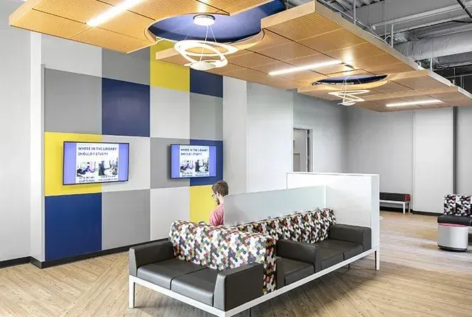 Modern waiting area with gray sofa, TVs, and patterned wall panels. A person sits on the sofa.