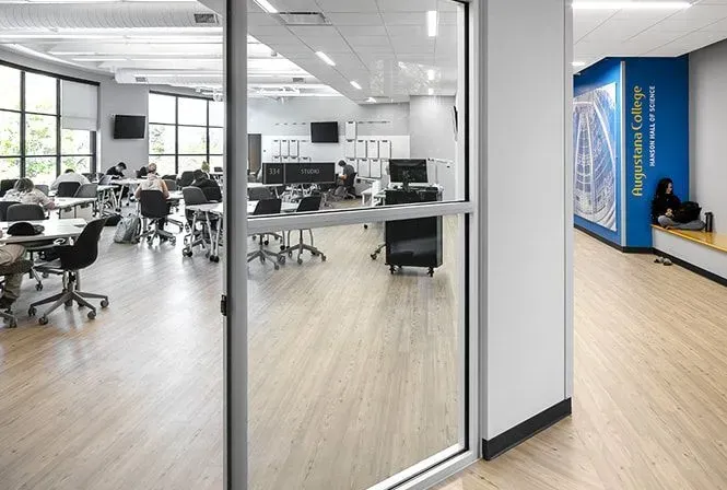 A classroom with students working on laptops. The room has large windows, light wood floors, and a blue wall.