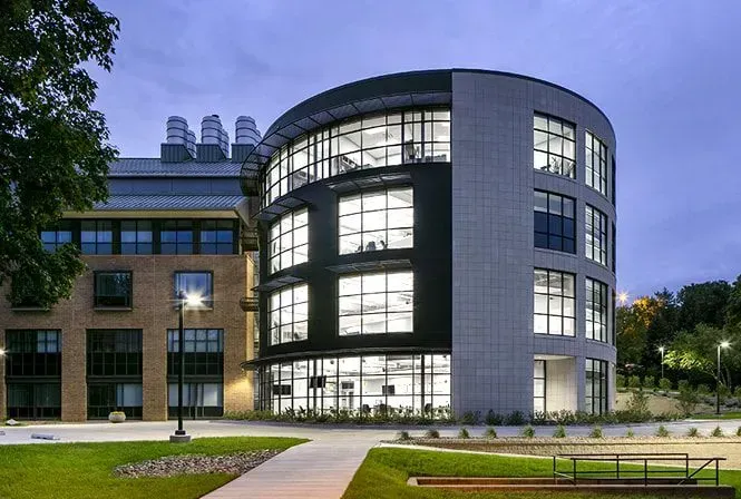 Modern, multi-story building with a rounded glass facade, adjacent to a brick building, on a green lawn.