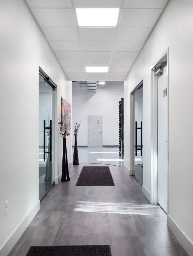Bright hallway with white walls and gray flooring, leading to a door at the end.  Dark accents include mats and vases.