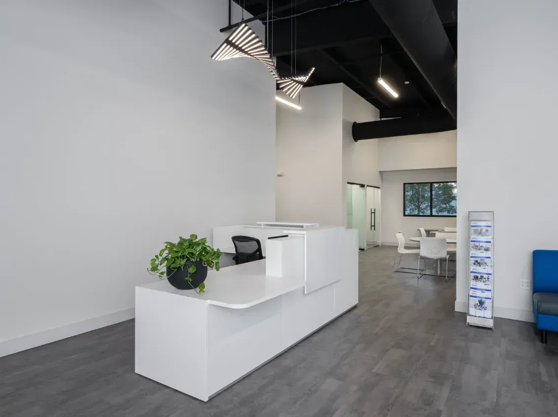 Modern white reception desk in an office with gray flooring, a plant, and a waiting area.