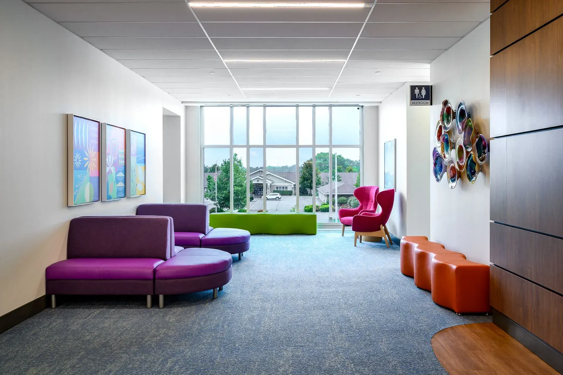 Hallway with seating and large windows. Purple, orange, and red furniture, artwork on walls, and a view of trees outside.