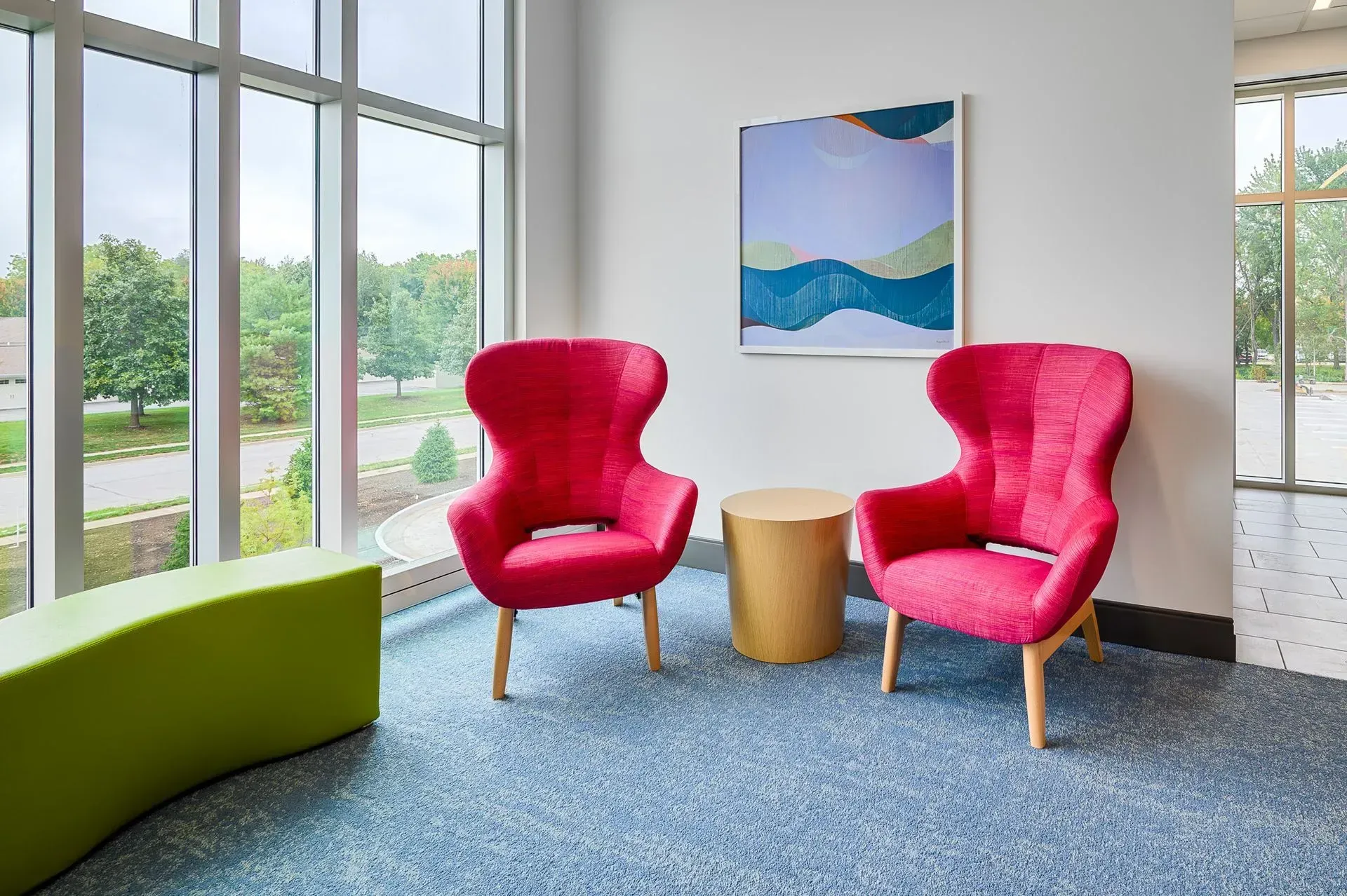 Two pink armchairs and a small round table in front of a window, artwork on the wall.