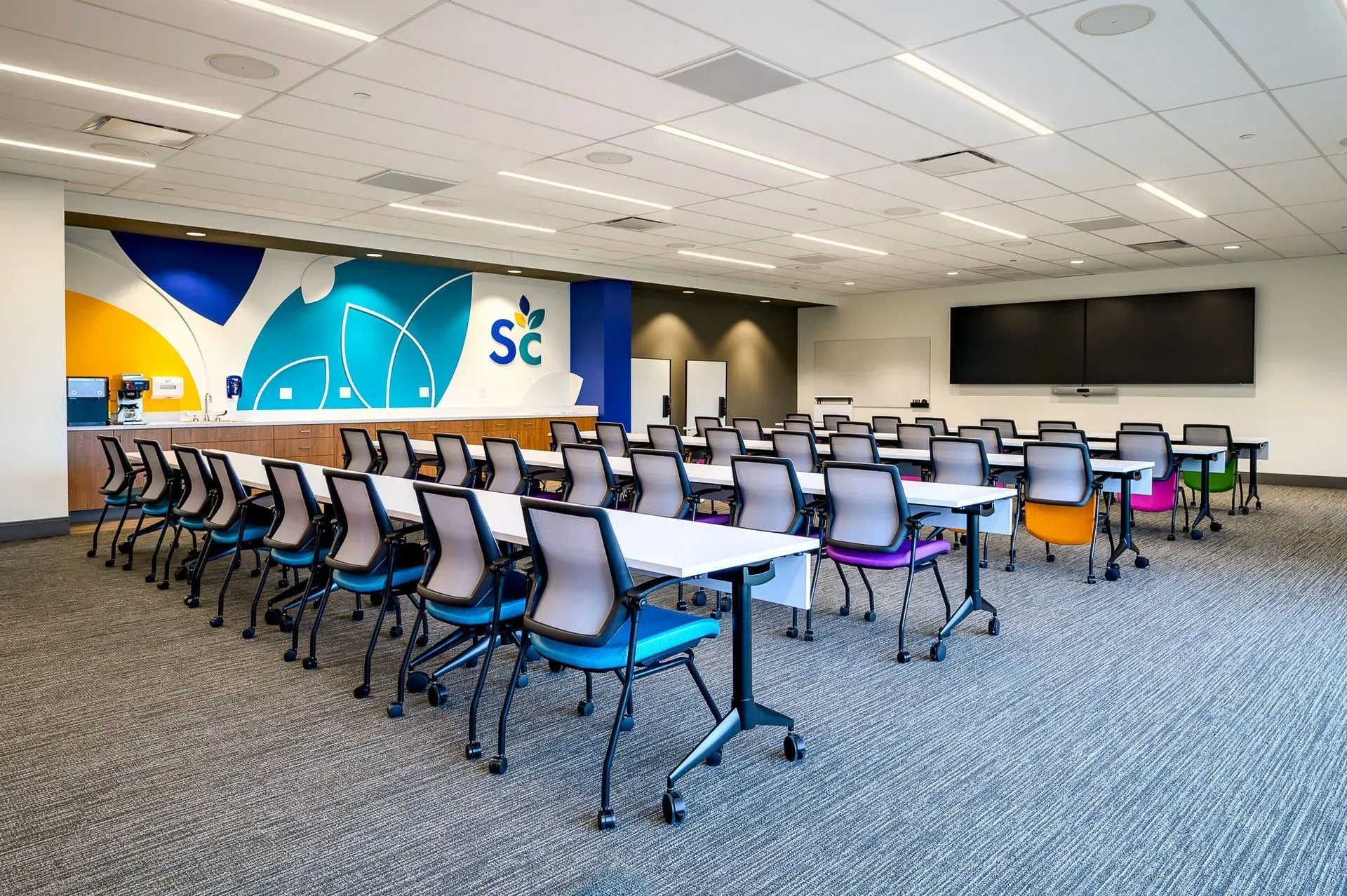 Classroom with rows of white tables and colorful chairs, large screen, and logo on wall.