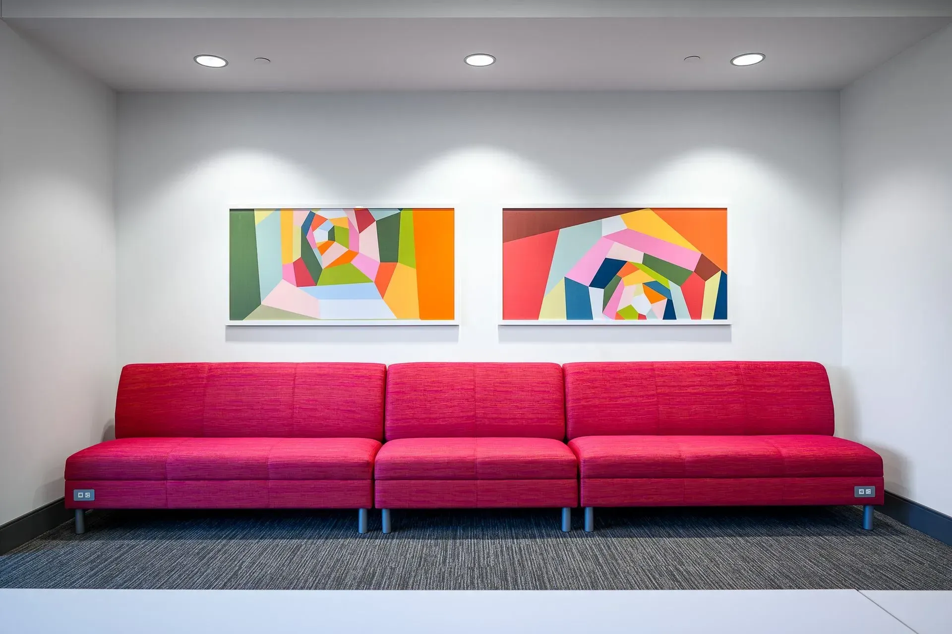 Red modular sofa beneath two colorful abstract paintings, in a white-walled room.