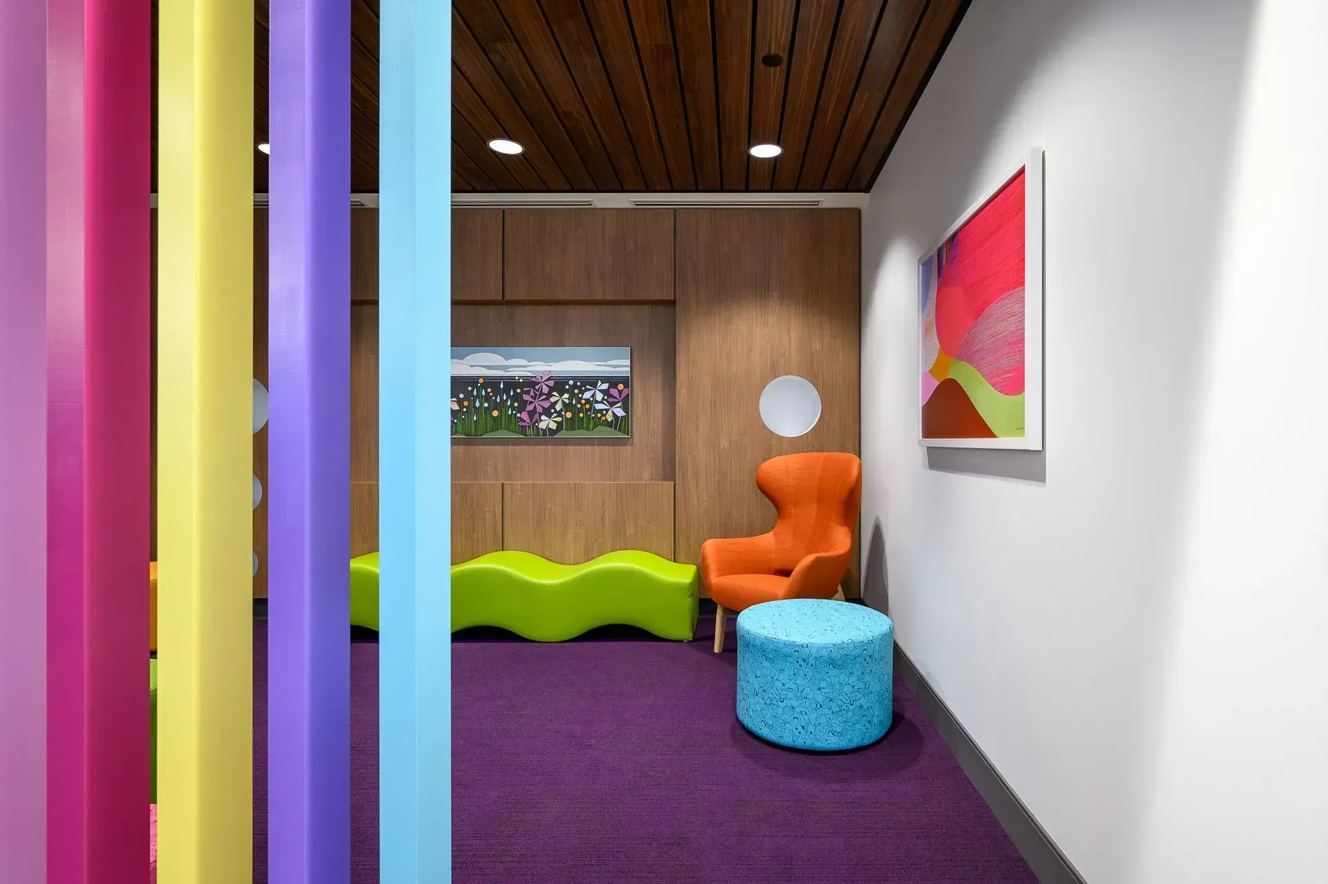 Colorful waiting room with purple carpet, orange chair, and artwork. Wooden accents and bright pillars.
