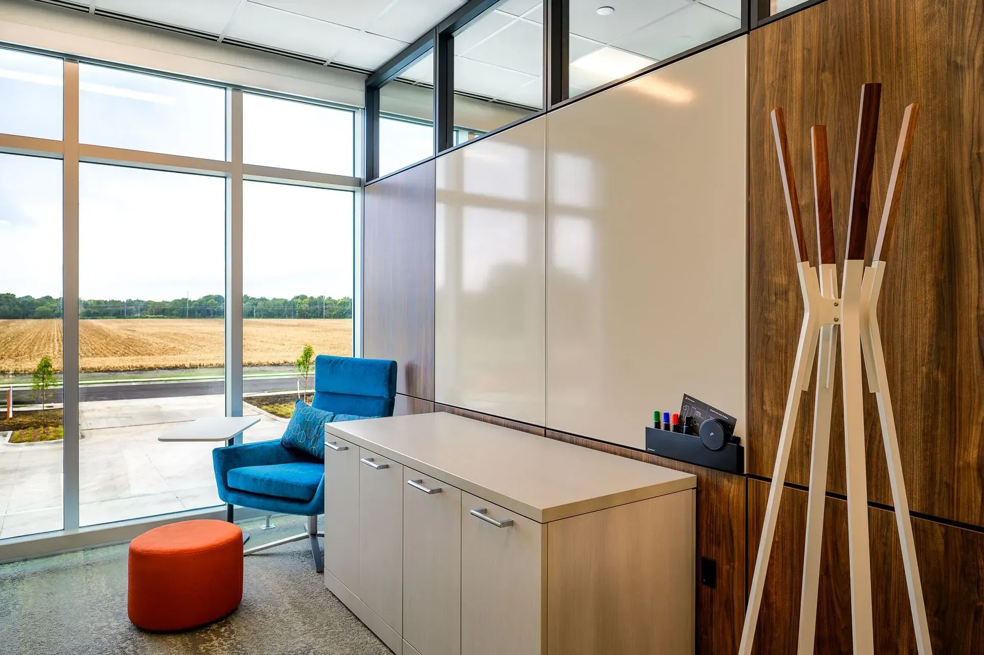 Office space with large windows, blue chair, orange ottoman, whiteboard, coat rack, and cream storage cabinet.