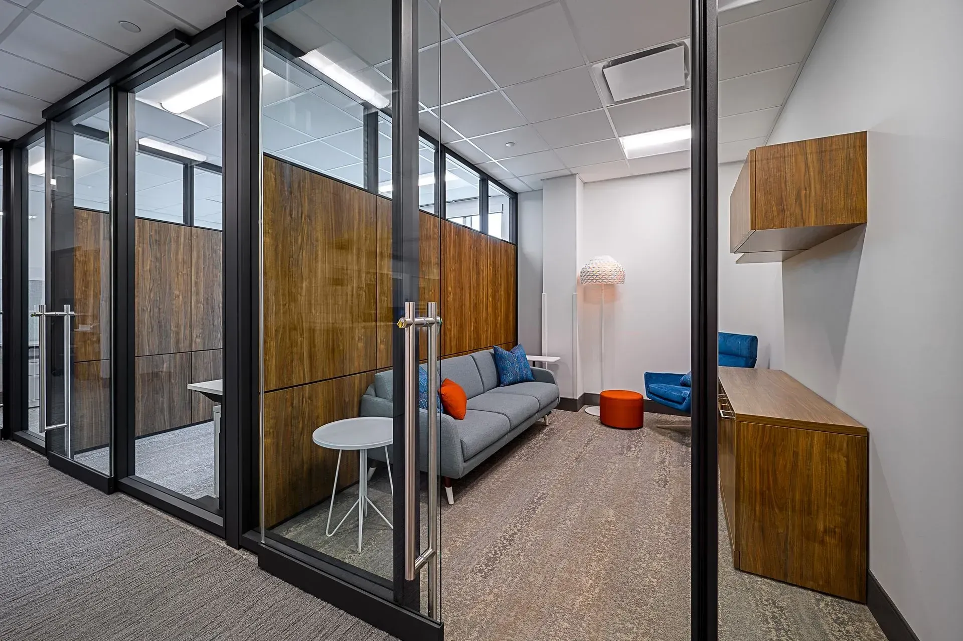 Modern office interior with glass-walled rooms. One room has a sofa, chairs, and a small table.