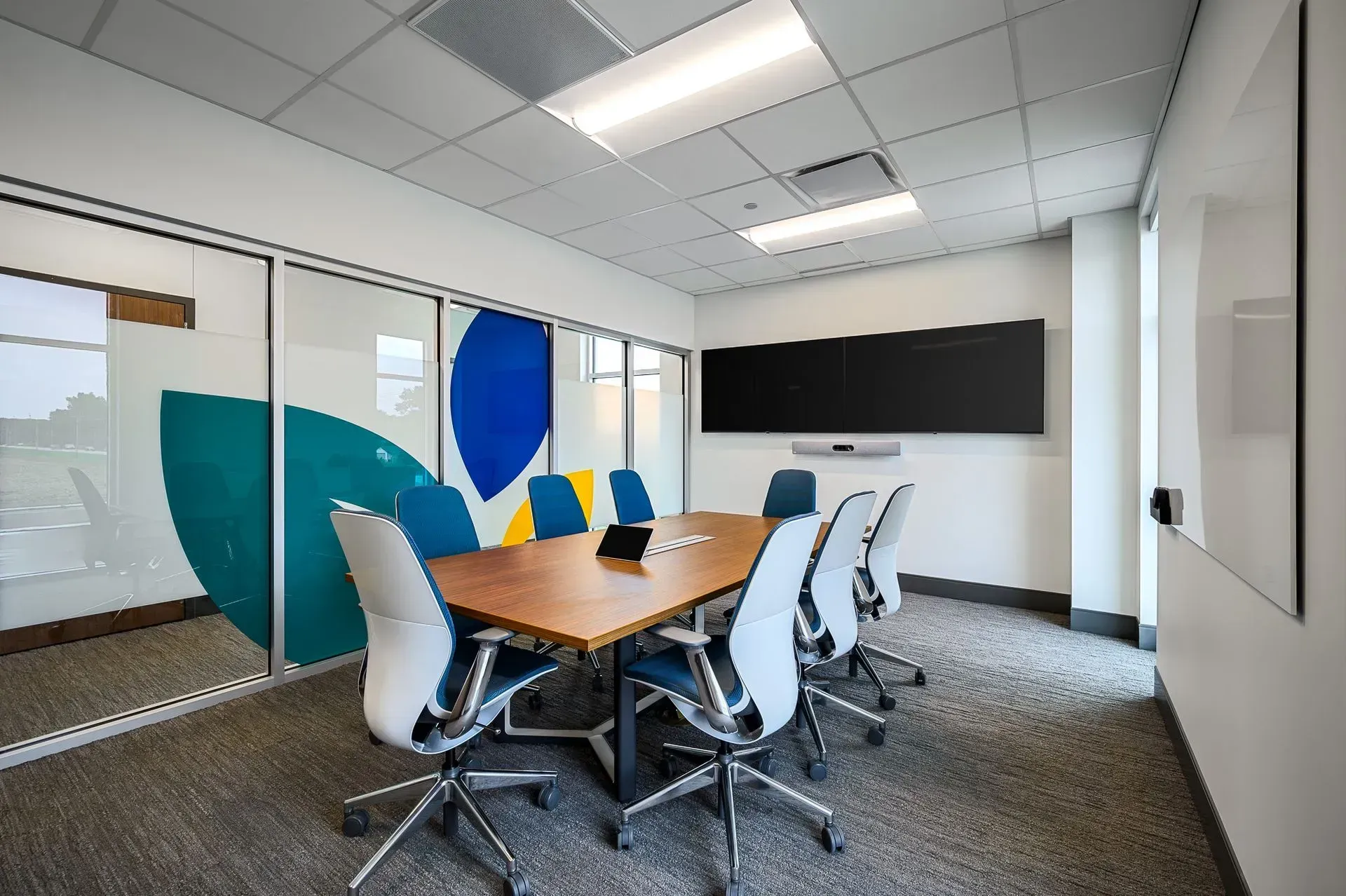 Conference room with wooden table, blue/yellow chairs, large screen, glass wall with blue and teal shapes.