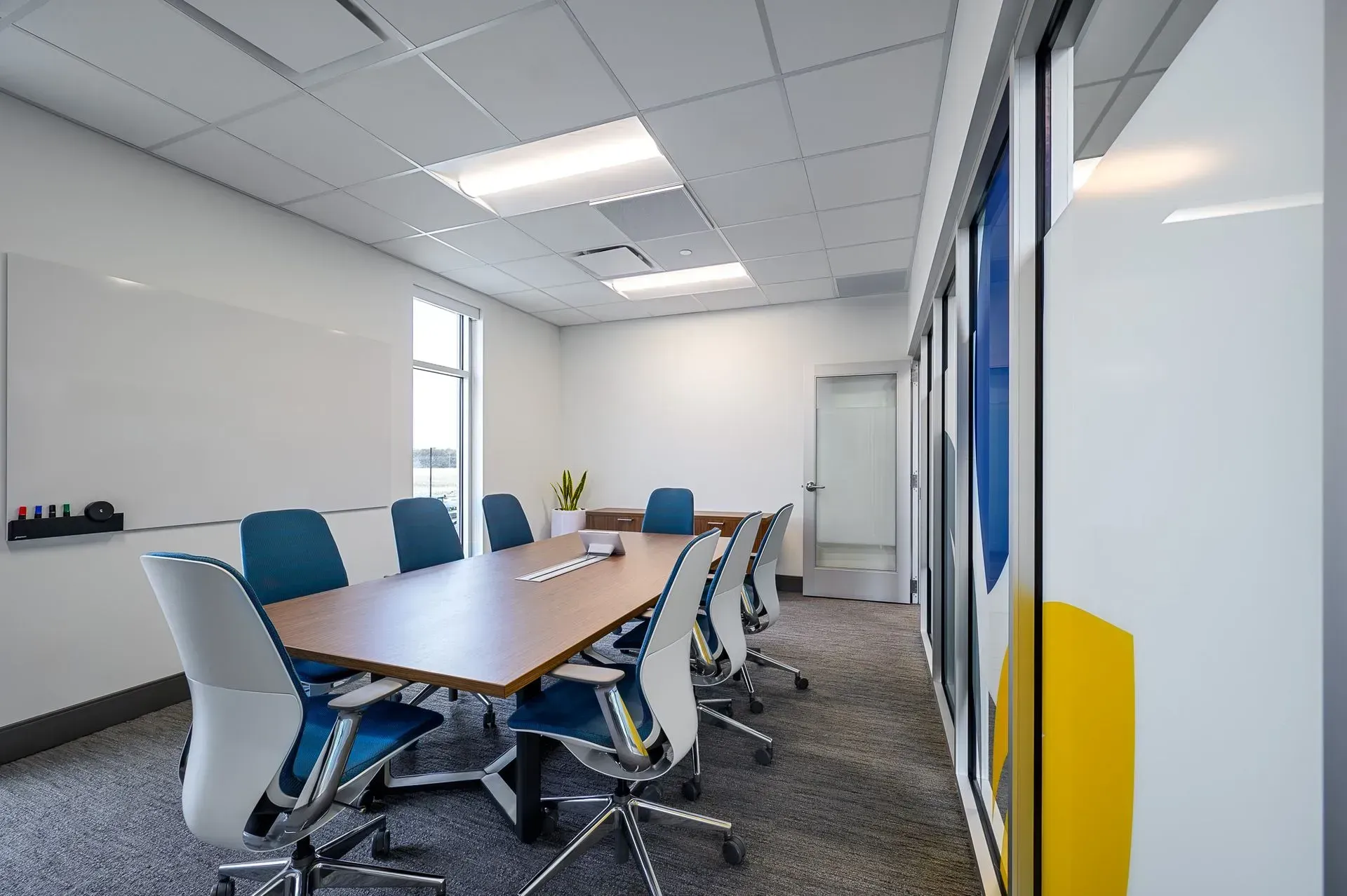 Conference room with a long table, chairs, whiteboard, and window. Blue and white color scheme.