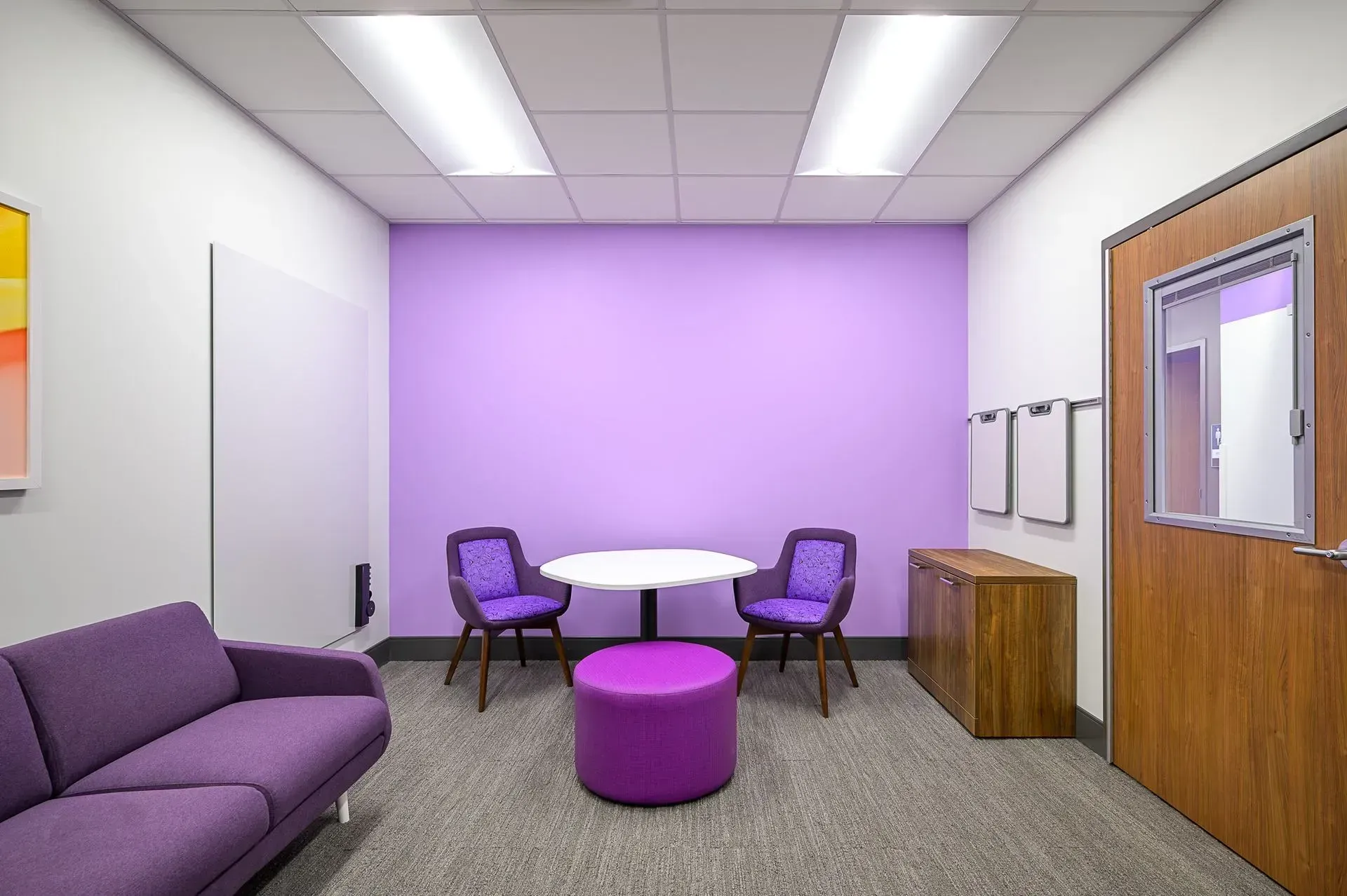 Purple-themed waiting room with sofa, chairs, round table, and a wooden cabinet next to a door with a window.