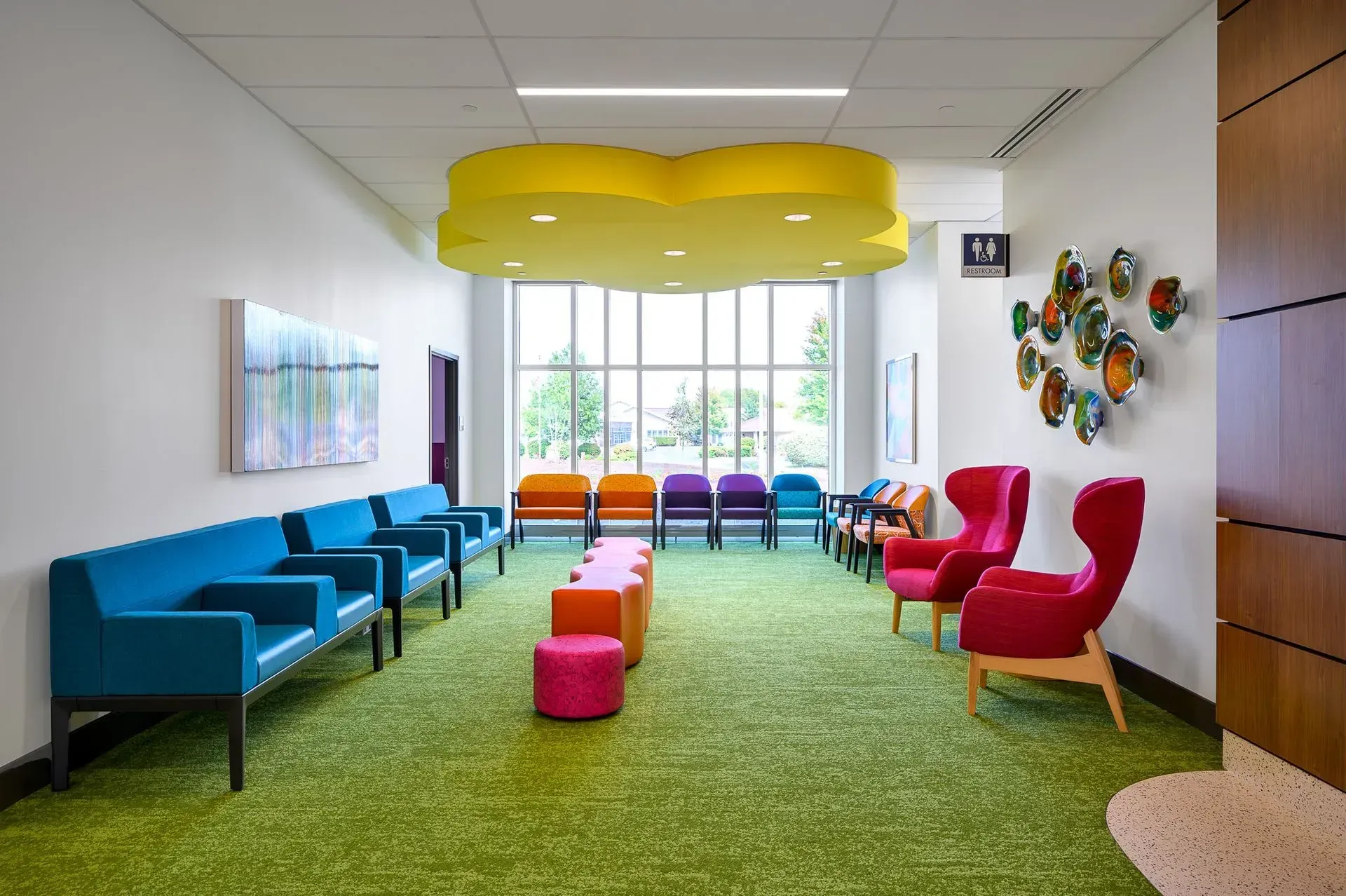 A colorful children's waiting room. Bright green carpet, various colored chairs, whimsical ceiling light, and artwork.