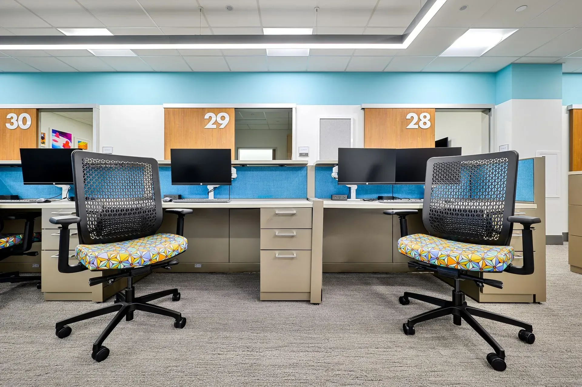 Office cubicles with desks, monitors, and chairs. Blue and white walls, numbered cubicle dividers.