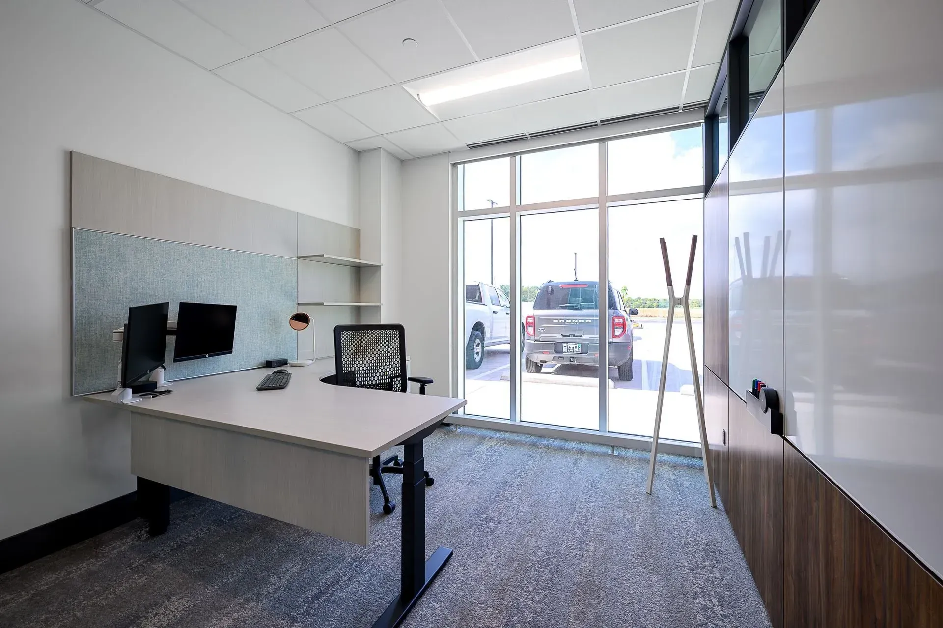 Modern office with desk, computer monitors, and large windows looking out onto a parking area.