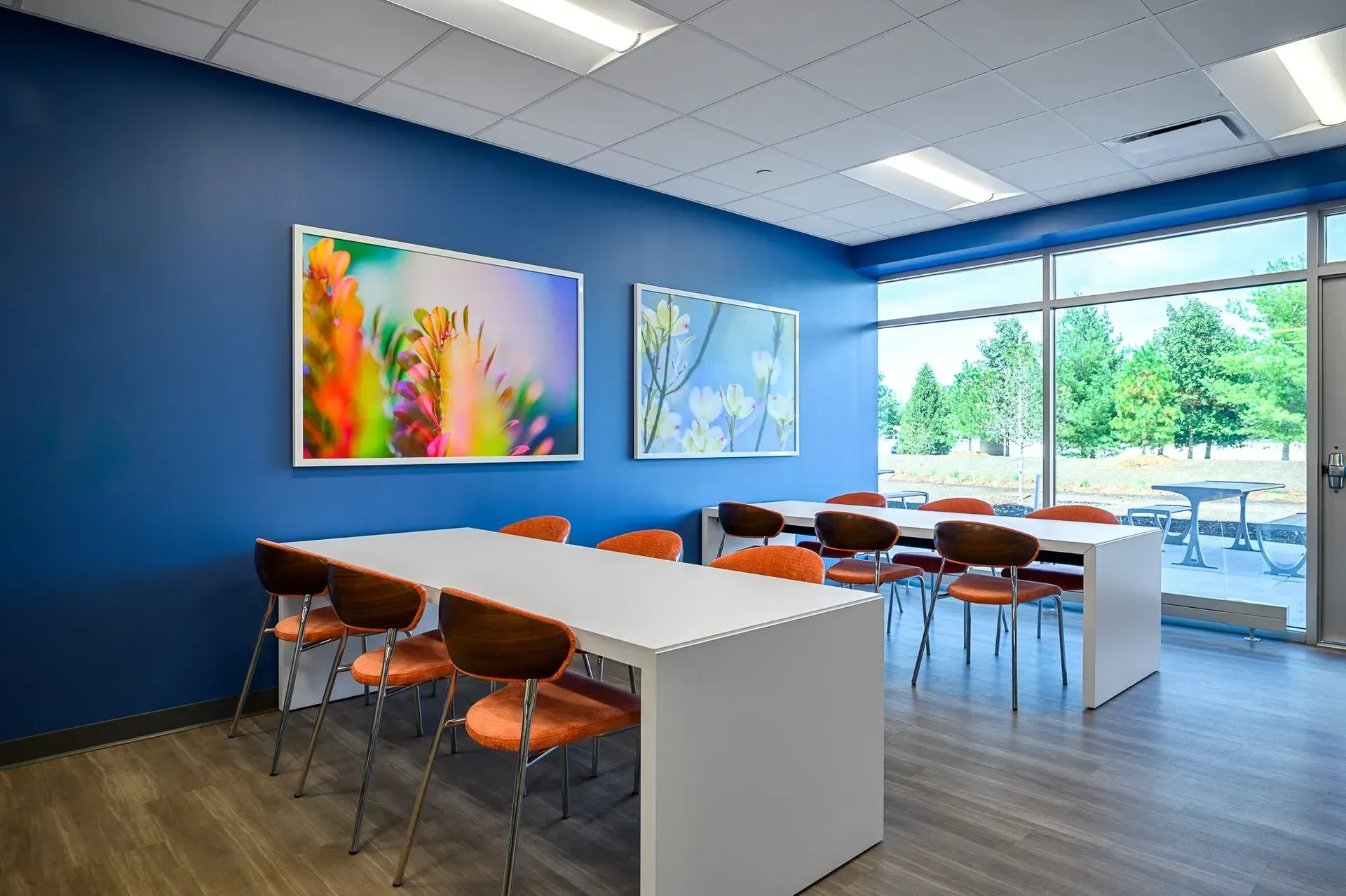 A modern room with tables, orange chairs, and art on a blue wall. Large windows reveal an outdoor patio.