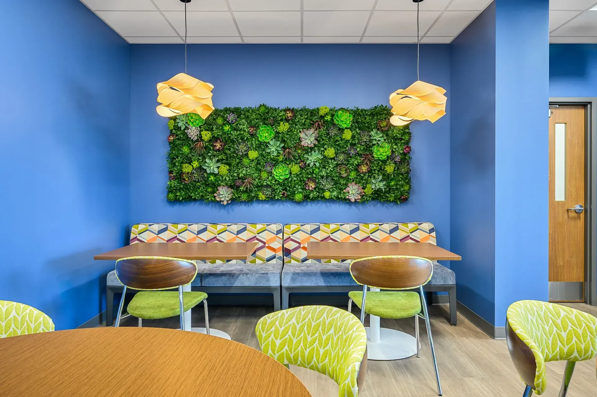 Cozy seating area with blue walls, a green plant wall, and two patterned benches. Wooden tables and lime-green chairs.