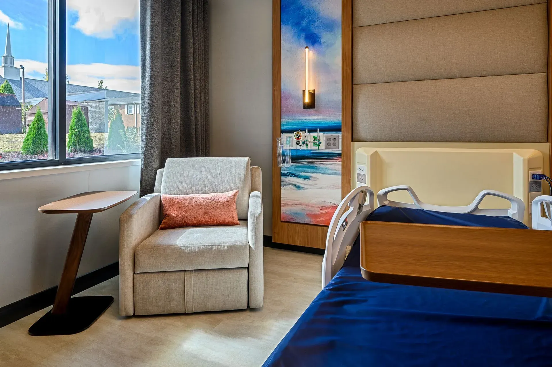 Hospital room with a window, chair, and patient bed. Bright, with a painting on the wall.