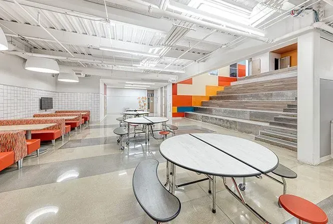 School cafeteria with orange booths, round tables, and tiered seating with colorful accents.