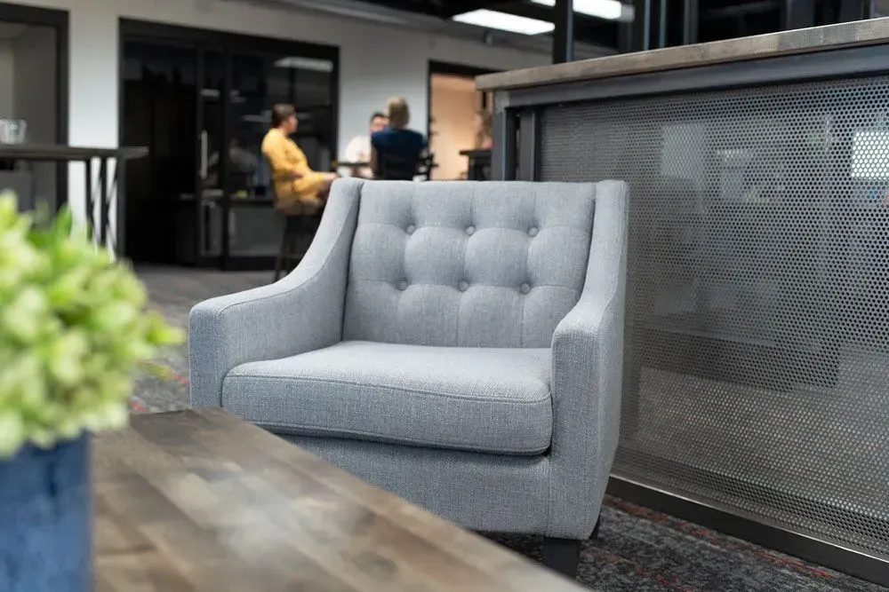 Gray upholstered armchair in a modern office space, with other furniture and people in the background.