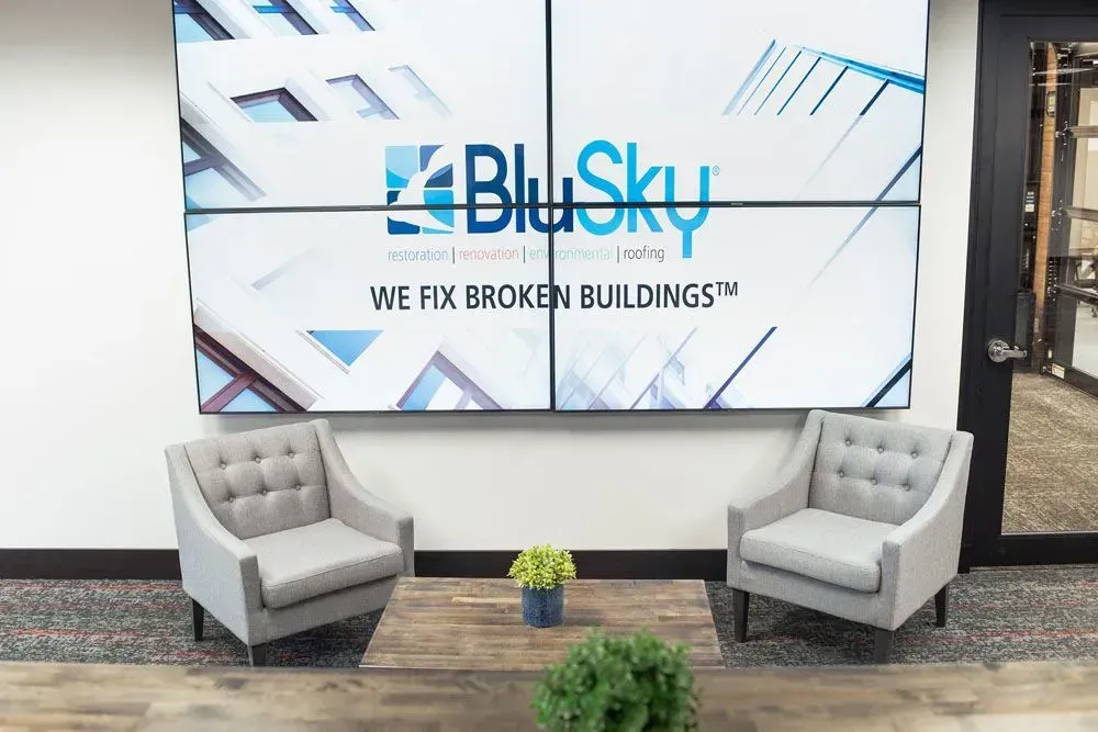 Two gray armchairs face a table with a plant, beneath a wall display with BluSky logo.