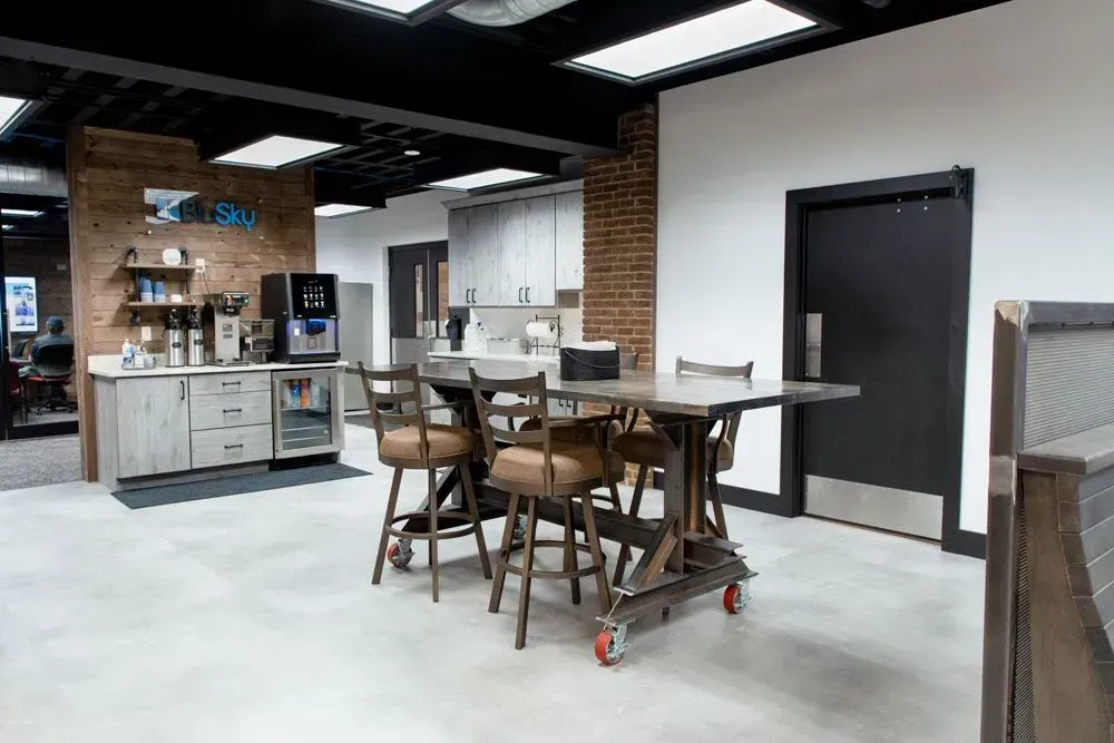 Office break room with industrial table and chairs, brick accent wall, coffee station, and kitchen.