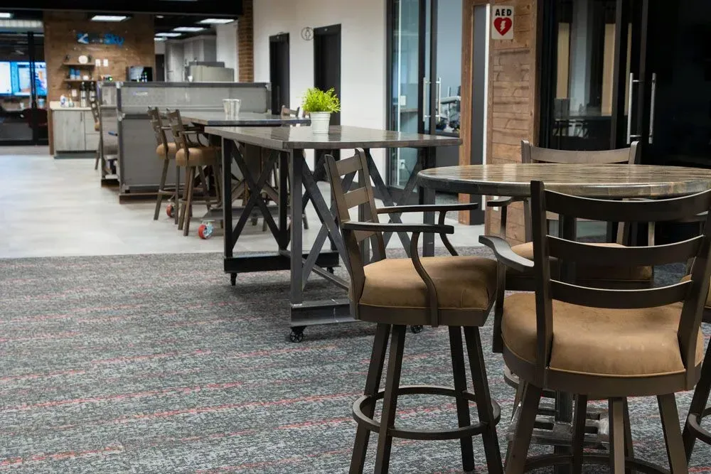 Office breakroom with tables, stools, carpet, and a brick accent wall.