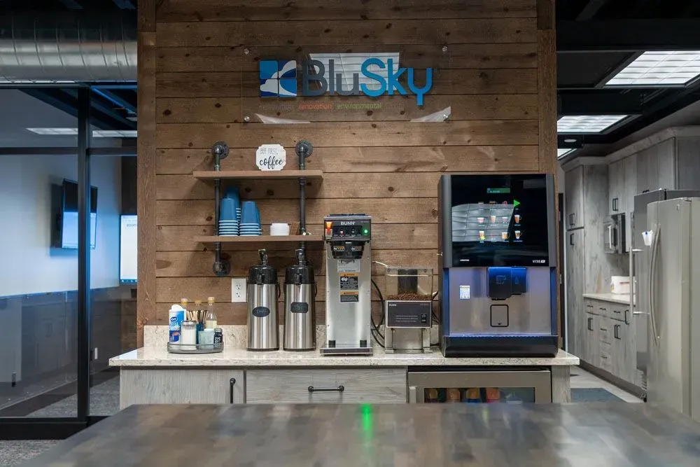 Coffee station in an office kitchen with espresso machines, syrups, and the BlueSky logo on a wood-paneled wall.
