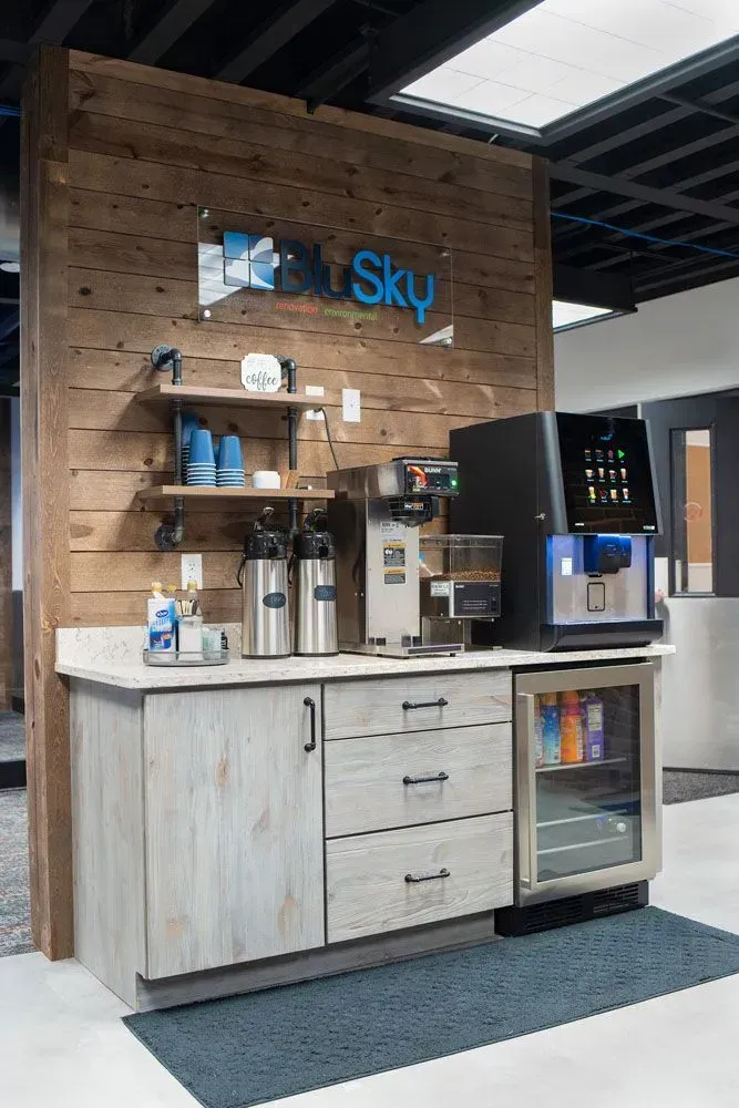 Coffee station in an office: wooden wall, coffee machines, fridge, cabinets, and a blue logo.