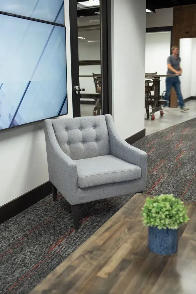 Gray armchair in an office setting. A plant and a wooden table are in front. A person is walking in the background.