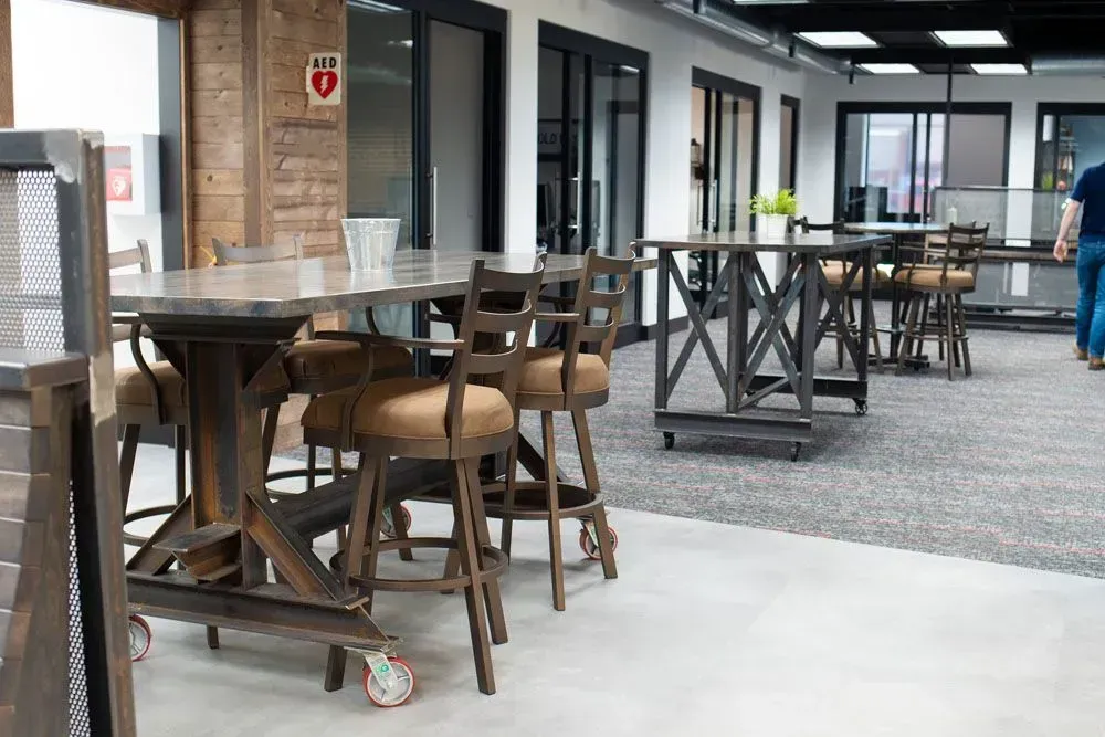Office breakroom with industrial-style tables and stools on wheels; white walls and carpet.