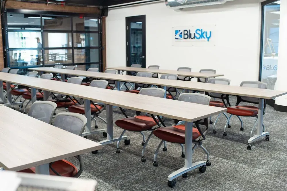 Classroom with long tables, chairs, and BluSky logo on the wall. The room has a glass door and carpet.