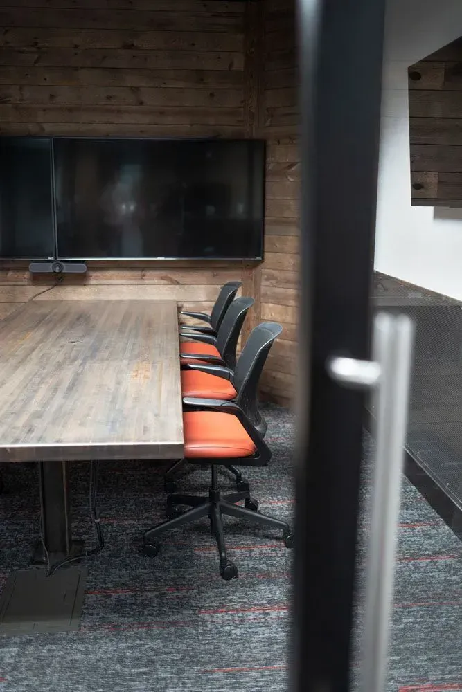Conference room with a long table, orange and black chairs, and a large TV on a wood-paneled wall.