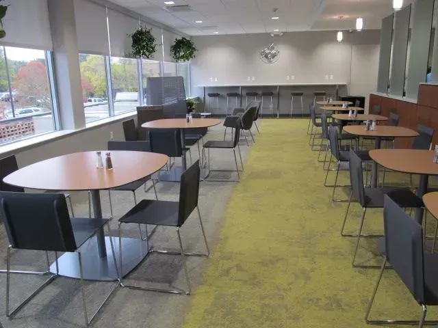 Cafeteria with round tables and chairs.  Large windows, bar seating, green carpet, and hanging plants.