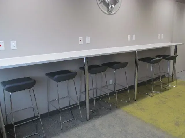Long white countertop with silver legs and five black bar stools against a light gray wall.