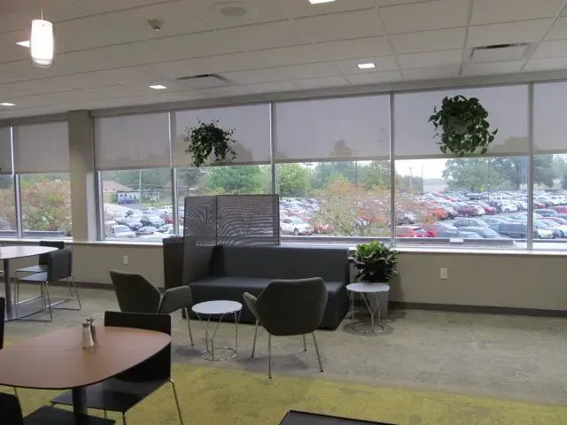 Interior lounge with seating, large windows, and plants. Parking lot visible outside.