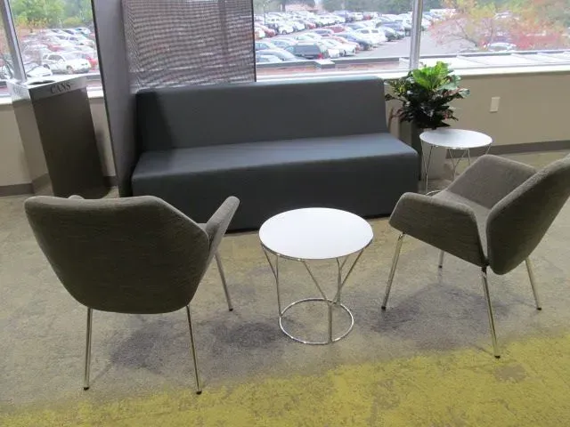 Lounge area with gray sofa, two armchairs, and small white tables. Windows overlook parking lot.