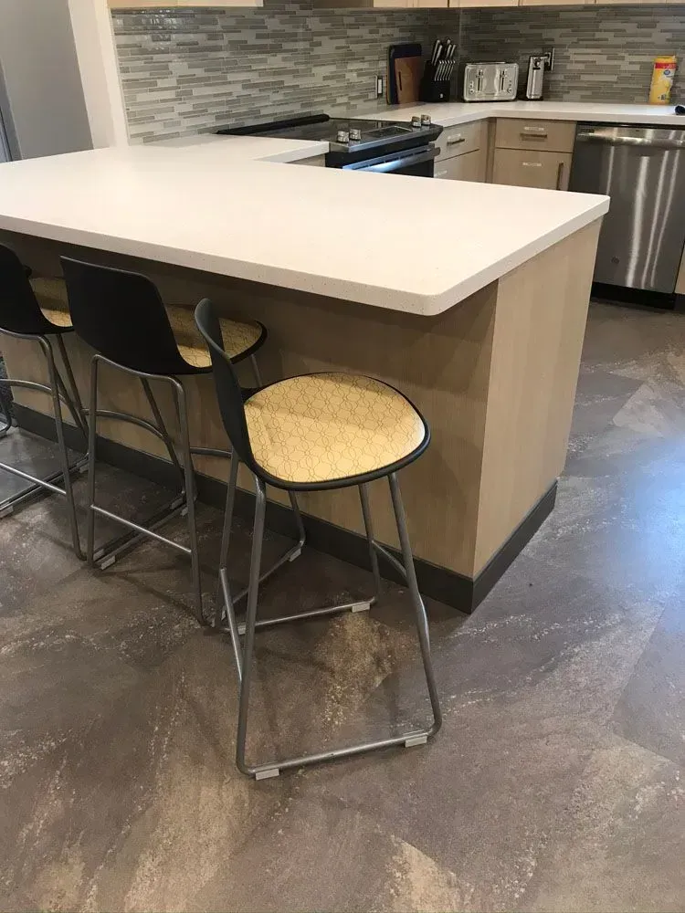 Kitchen island with white countertop, three black and tan barstools, stainless steel appliances, and brown cabinet base.