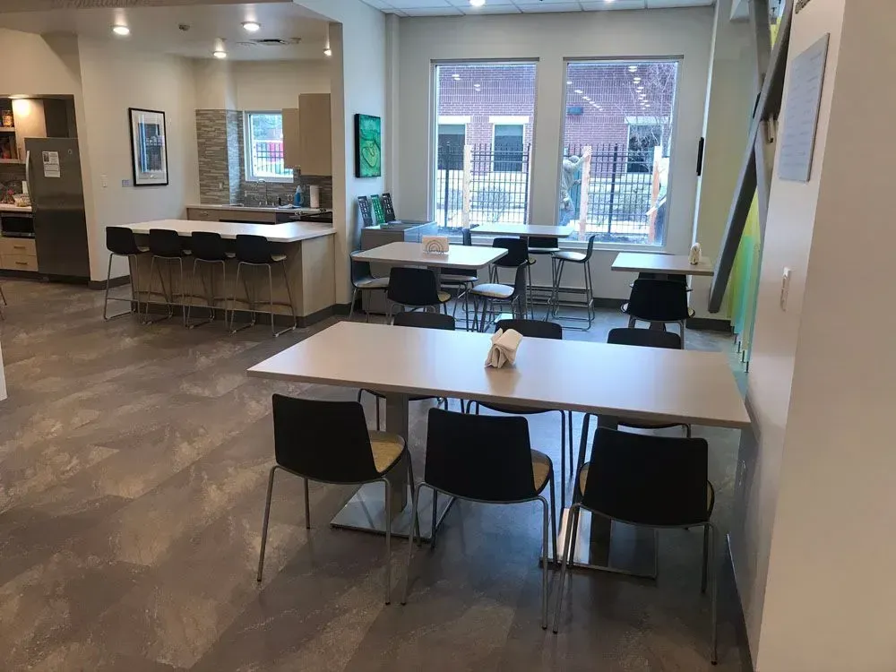 Open office breakroom with tables, chairs, and a kitchen area with stools and appliances. Gray floors and neutral walls.