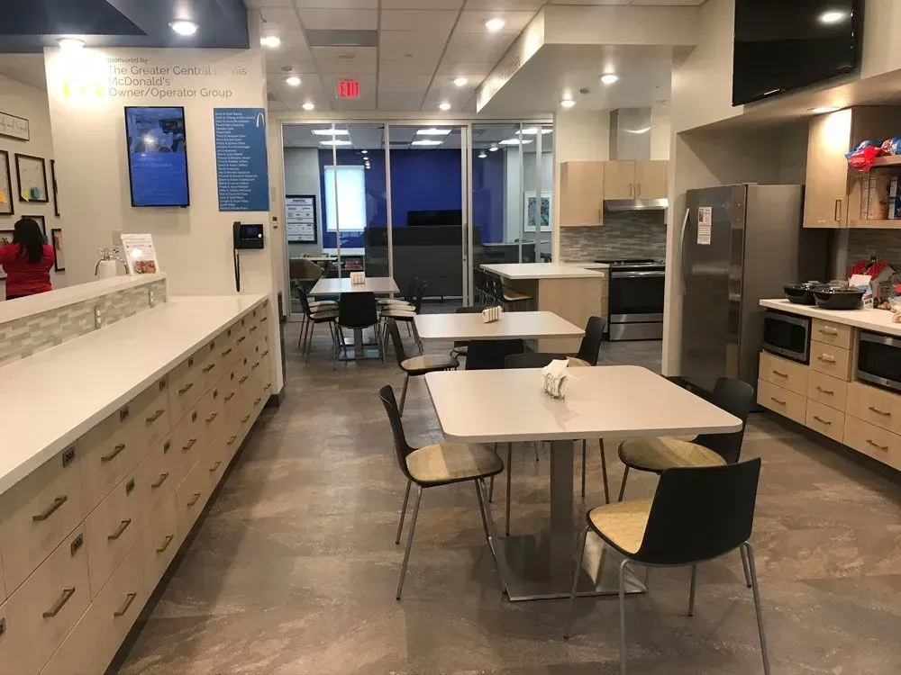 A modern office break room with tables, chairs, and a kitchen. Bright lights and neutral colors dominate the space.
