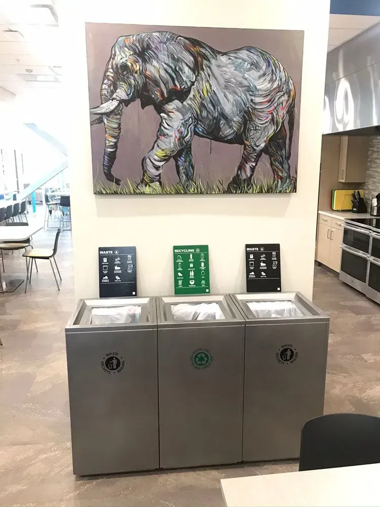 Three recycling bins beneath an elephant painting in a brightly lit room.