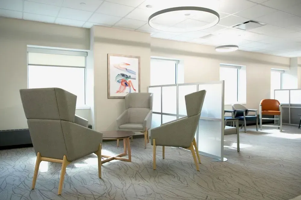 Waiting room with armchairs, table, artwork, windows, and partial cubicle walls.