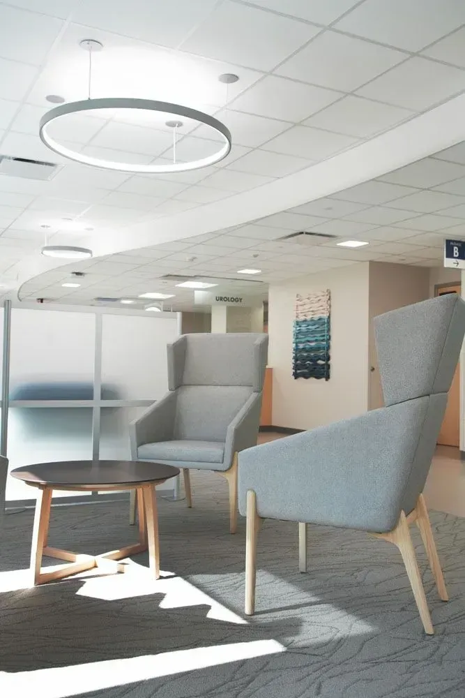 Lounge area with gray chairs, wooden side table, and modern circular light fixture; hallway with art in background.
